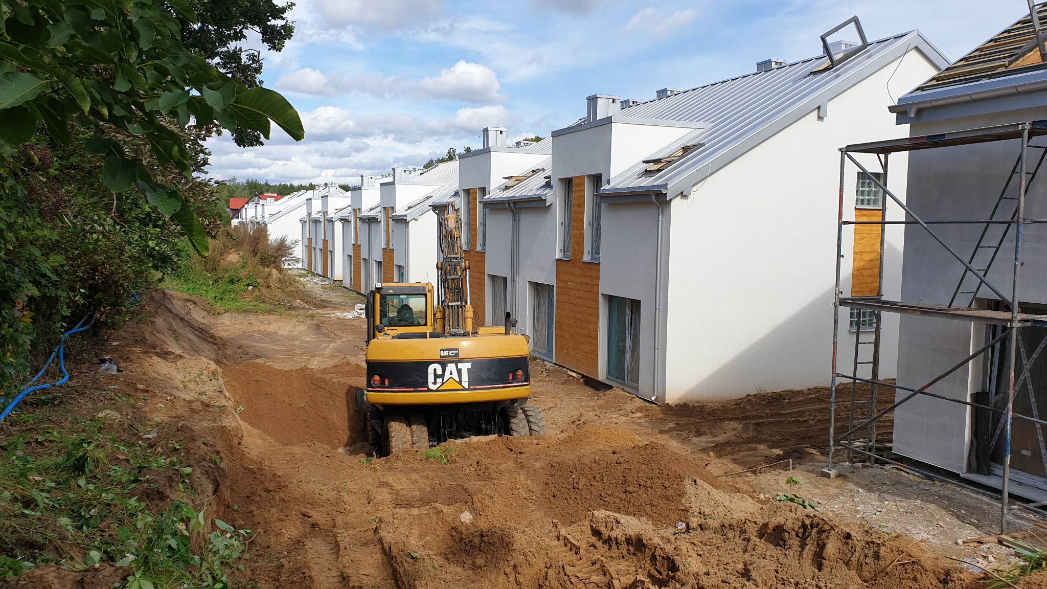 Żółta koparka CAT wykonuje prace ziemne przed szeregiem nowych, dwukondygnacyjnych domów z białymi elewacjami i szarymi dachami, częściowo jeszcze w rusztowaniach.