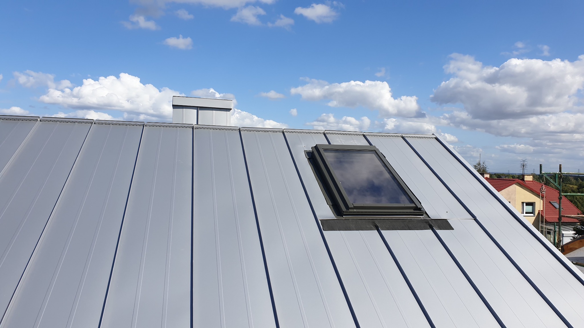 Szary dach z blachy trapezowej z oknem dachowym i kominem na tle błękitnego nieba z chmurami.