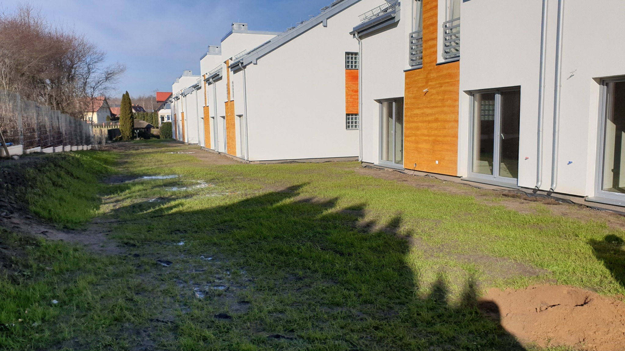 Szereg nowo wybudowanych, białych domów jednorodzinnych z drewnianymi akcentami, widocznych z perspektywy trawnika w trakcie zagospodarowywania terenu, z widocznymi śladami po pracach ziemnych.