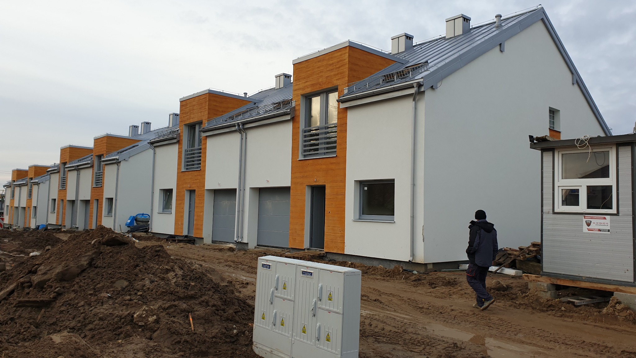 Rząd szeregowych domów w trakcie budowy z drewnianymi akcentami, widoczny niezagospodarowany teren i wędrująca osoba.