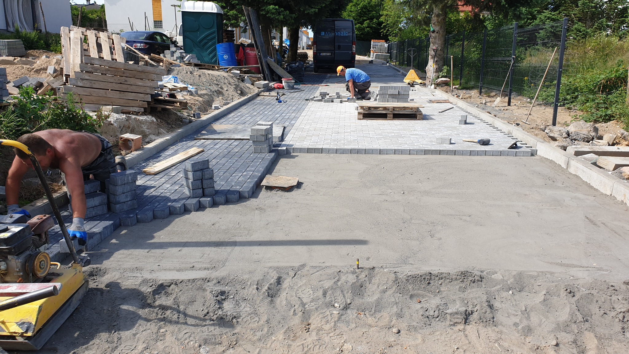 Układanie kostki brukowej na posesji: pracownik zagęszcza podłoże zagęszczarką, drugi układa ciemnoszarą kostkę, w tle ułożona jasnoszara kostka, widoczny przenośny WC i samochód dostawczy.