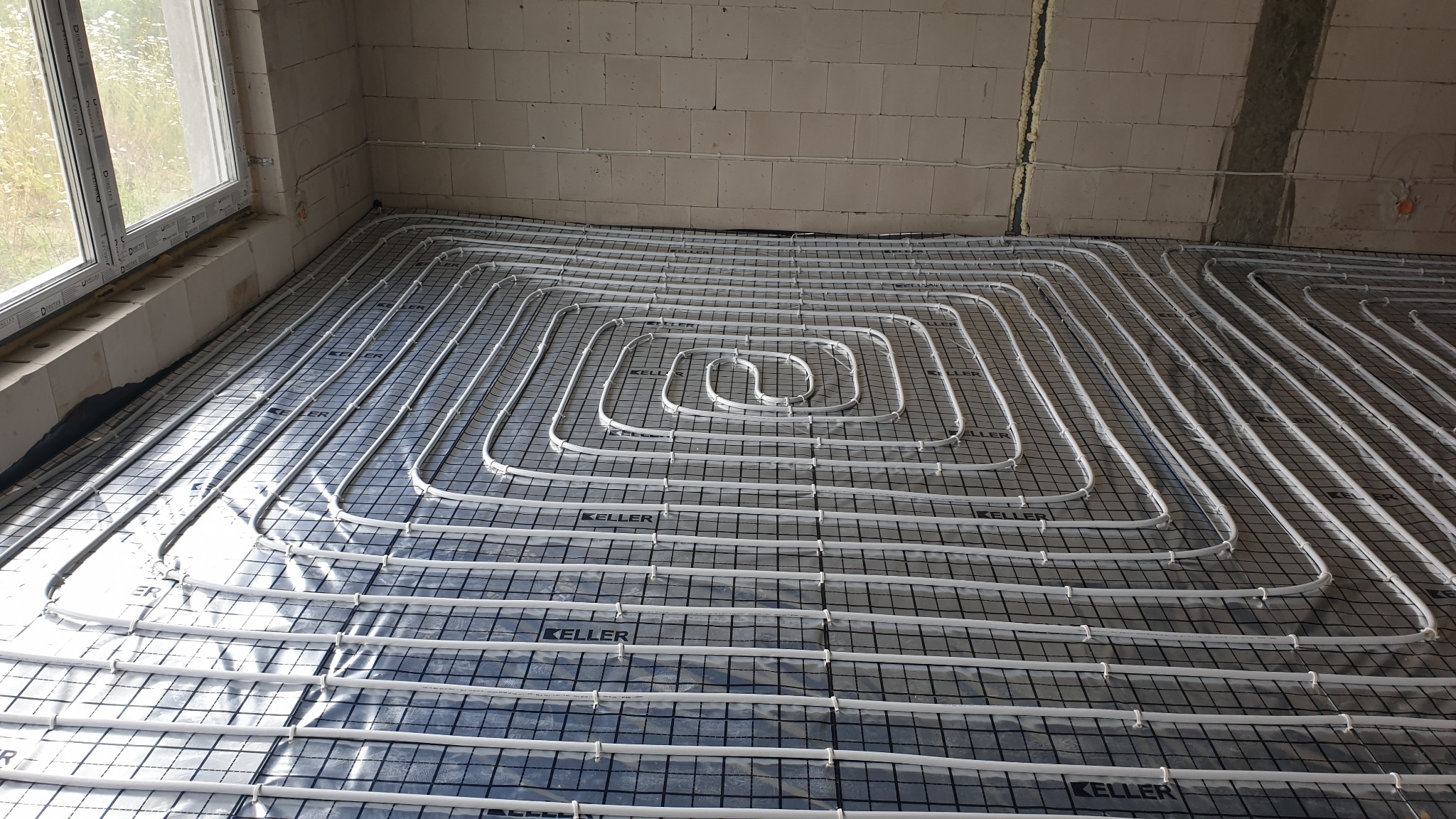 Montaż ogrzewania podłogowego w surowym stanie, widoczne białe rury ułożone w pętle na czarnej macie z siatką, ściany z białych bloczków.