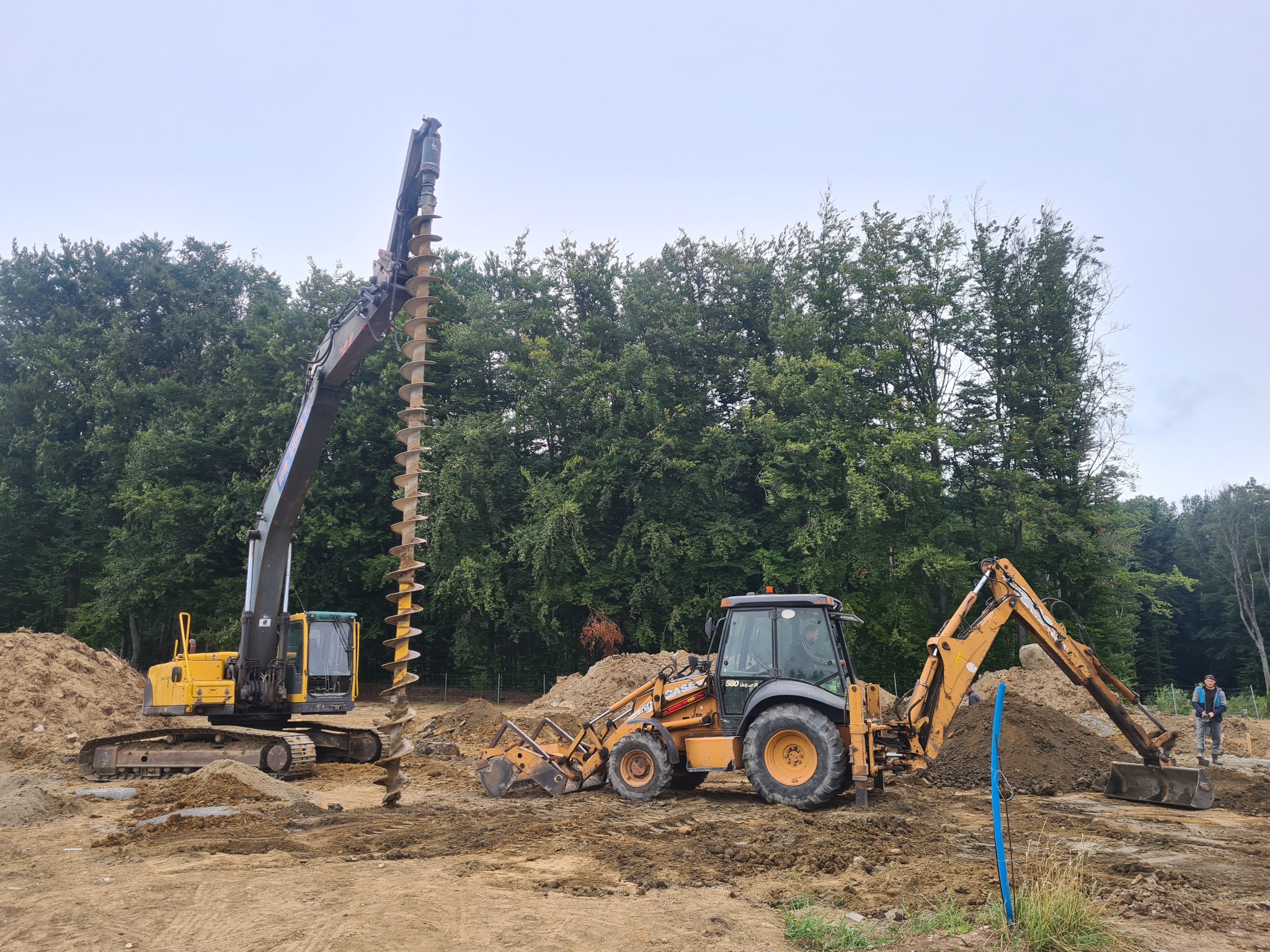 Żółta koparka z wiertłem ślimakowym i pomarańczowa koparko-ładowarka na placu budowy, w tle las i pochmurne niebo.