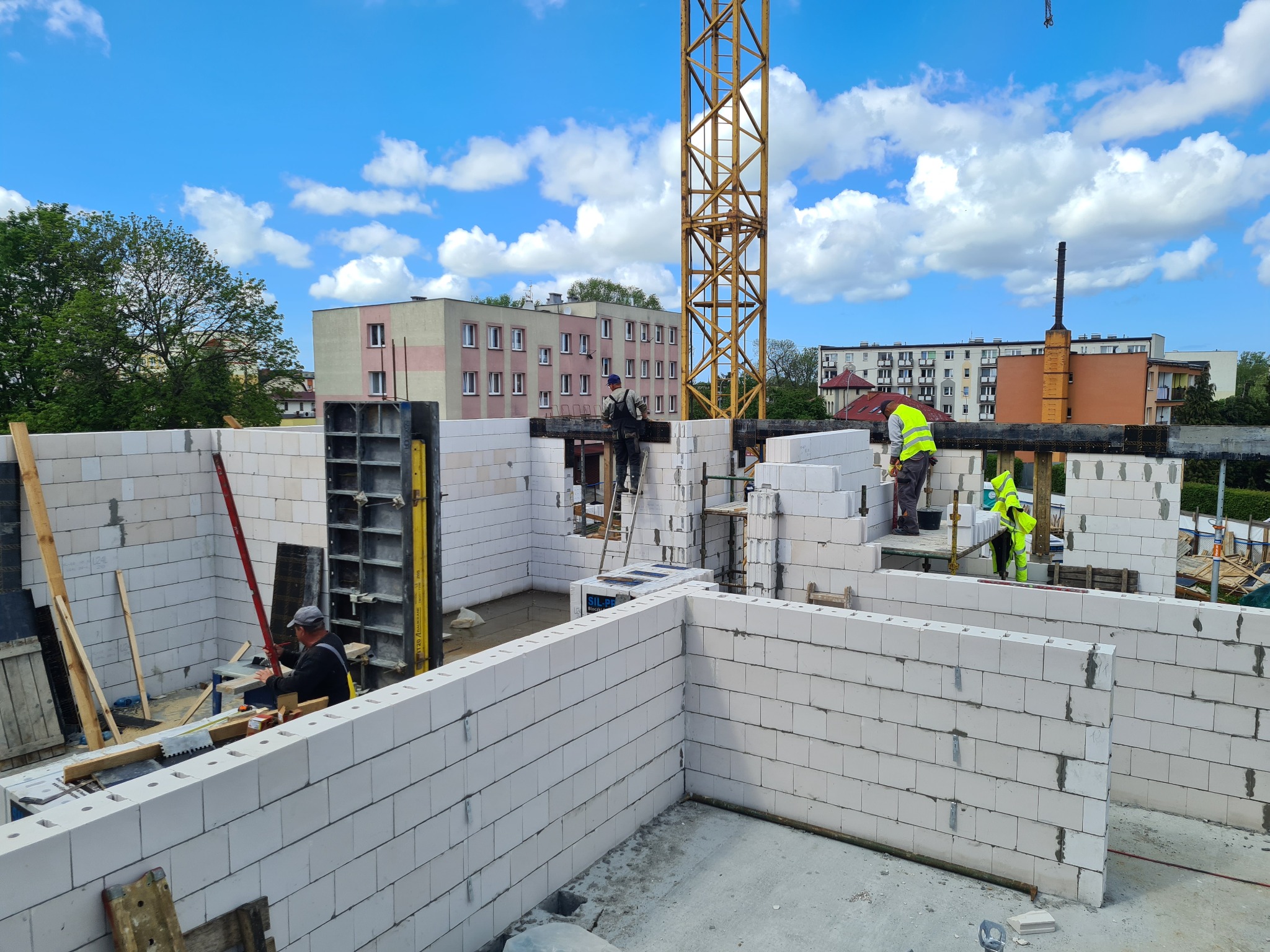 Budowa domu z białych bloczków: murarze w żółtych kamizelkach układają ściany, w tle żółty żuraw i budynki mieszkalne, błękitne niebo z chmurami.