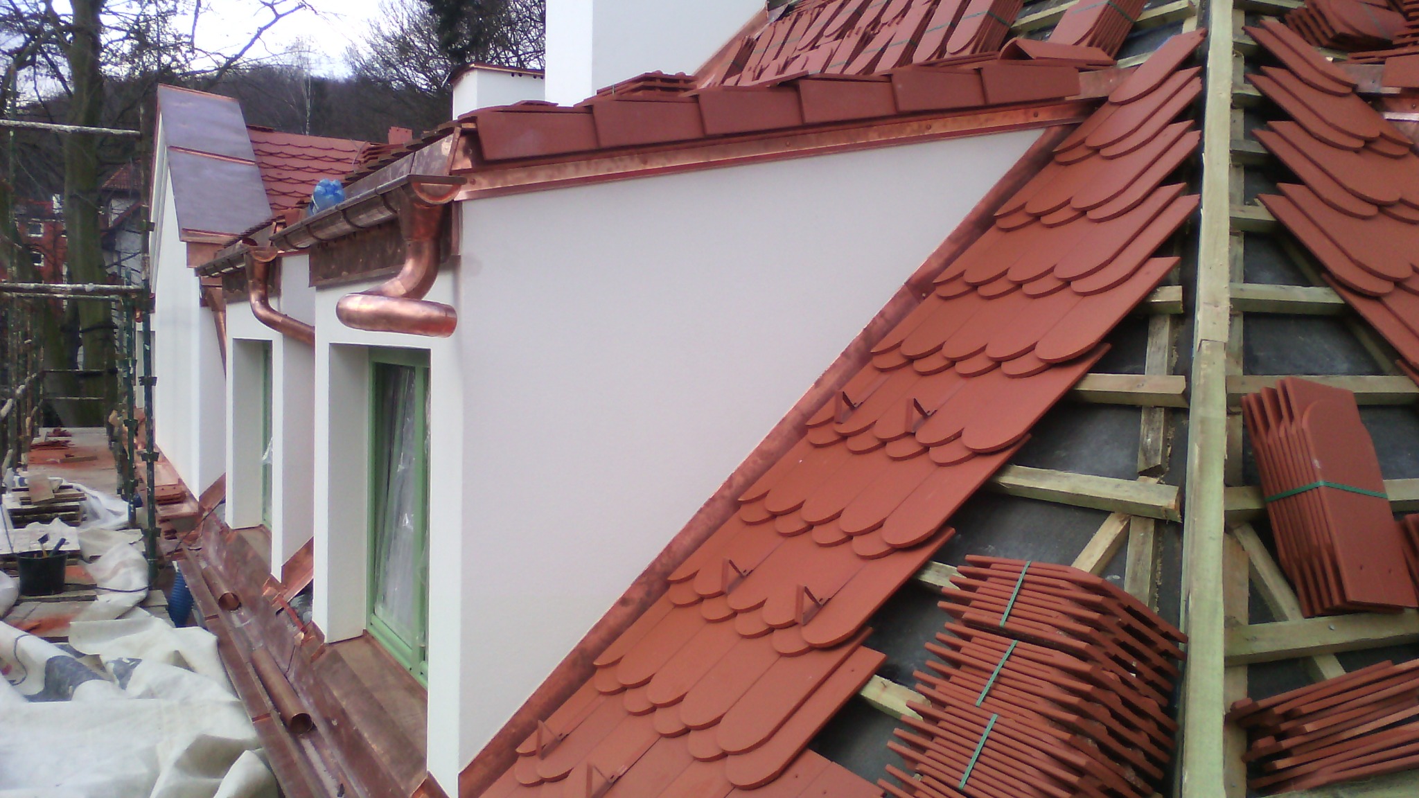 Montaż ceramicznej dachówki w kolorze ceglastym oraz miedzianych rynien na budynku z białymi ścianami, widoczne rusztowania i przygotowane paczki dachówek.