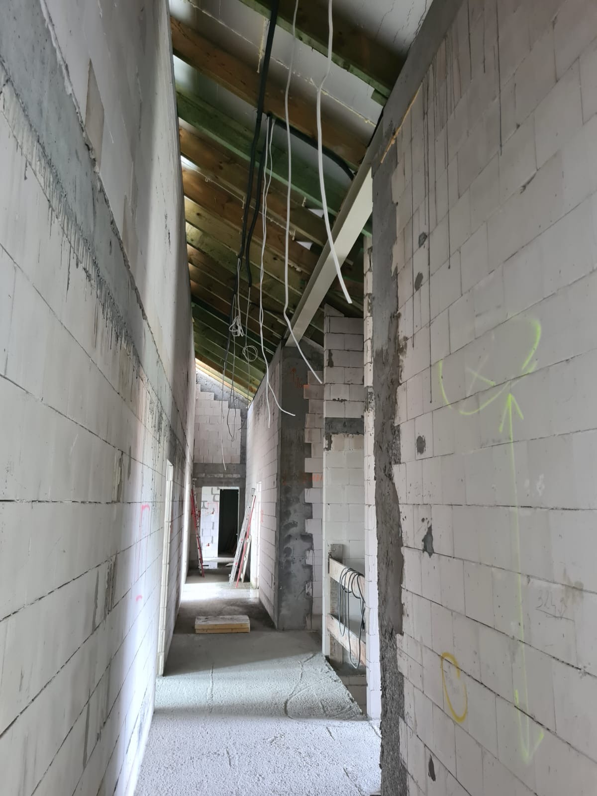 Wnętrze budynku w trakcie budowy: surowe ściany z białych bloczków, drewniany strop z widocznymi kablami, podłoga wylewka, pionowe strzałki narysowane na ścianie.