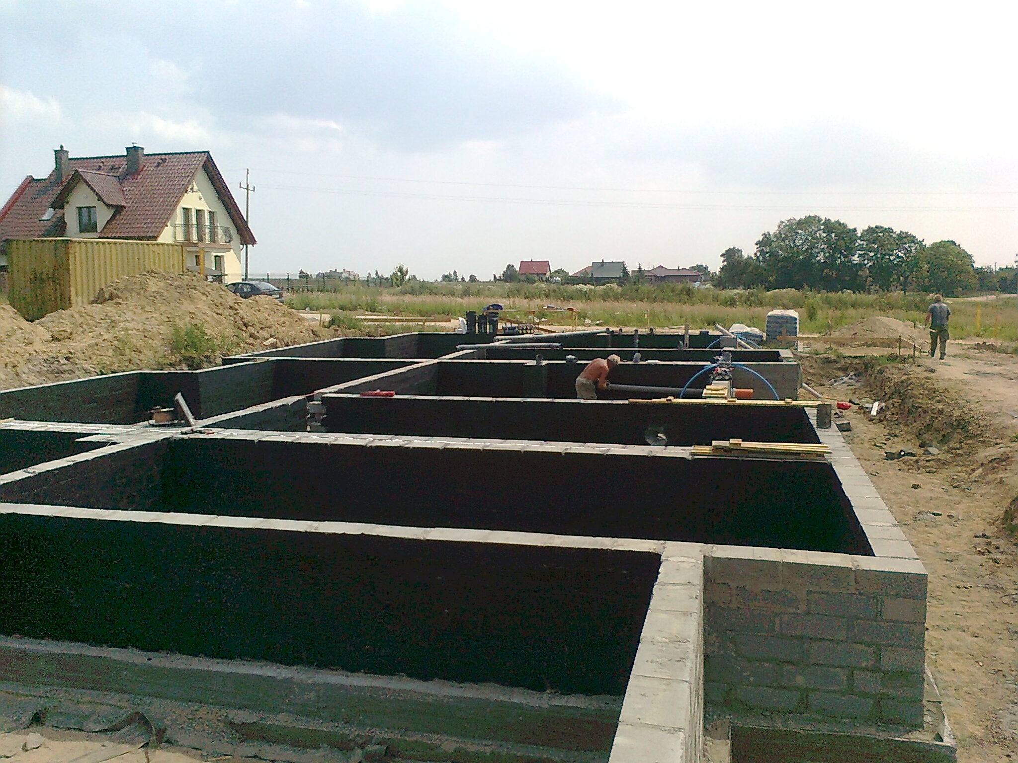 Fundamenty domu jednorodzinnego z hydroizolacją, widoczne zbrojenia i instalacje, w tle dom w budowie i krajobraz wiejski.