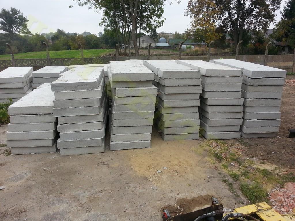 Stosy szarych, betonowych płyt chodnikowych ułożone na ziemi, widoczne lekkie zabrudzenia. W tle zieleń drzew i częściowo ogrodzenie.