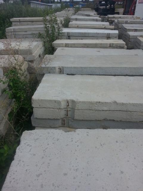 Stosy szarych, betonowych płyt prefabrykowanych z widocznymi elementami zbrojenia, ułożone w rzędach na zewnątrz, z roślinnością w tle.