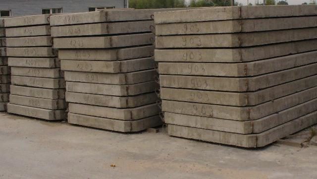 Stosy prefabrykatów betonowych ułożone jeden na drugim, widoczne numery seryjne na powierzchni płyt, szare tło, magazyn prefabrykatów.