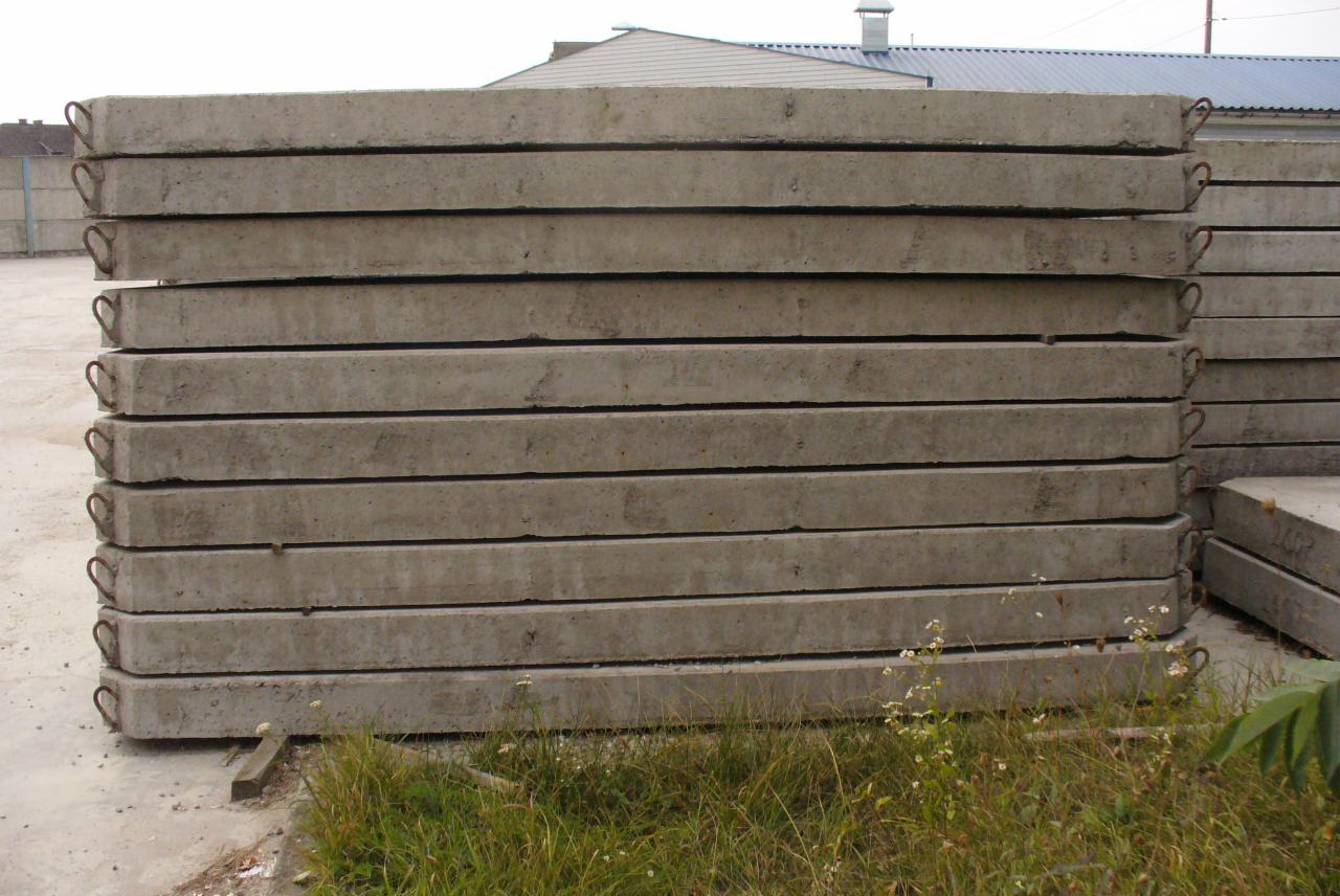 Stos prefabrykowanych płyt betonowych z uchwytami, ułożonych na zewnątrz, z widoczną trawą u podstawy i budynkiem w tle. Szare, surowe wykończenie.