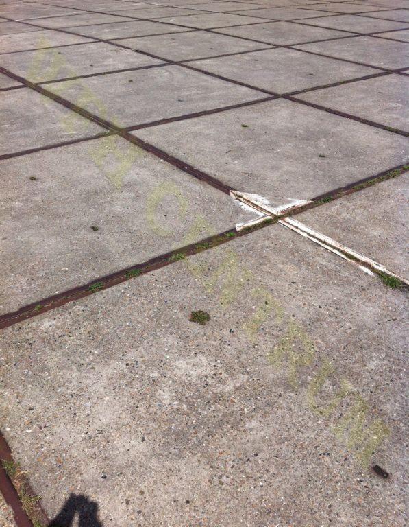 Betonowa nawierzchnia podzielona metalowymi listwami, z widocznymi śladami użytkowania i drobną roślinnością w szczelinach. Widoczny cień osoby w lewym dolnym rogu.