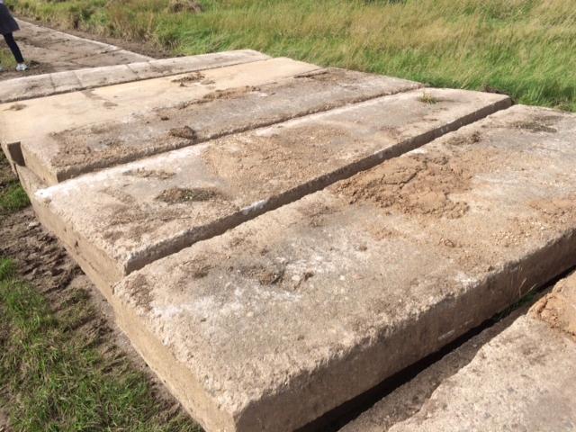 Ułożone, prostokątne płyty betonowe na trawiastym terenie, tworzące tymczasową drogę. Widoczne ślady użytkowania i zabrudzenia na powierzchni płyt.