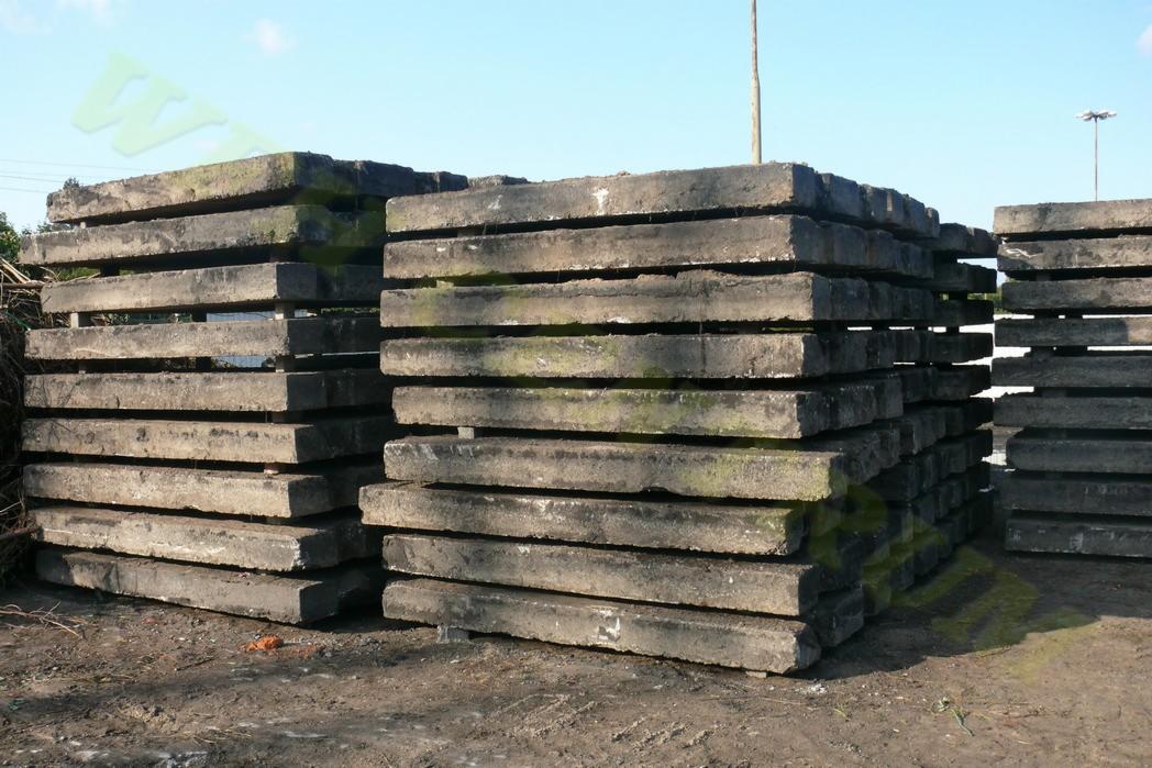 Stosy szarych, betonowych płyt chodnikowych ułożonych na ziemi, widoczne ślady użytkowania i mchu, w tle błękitne niebo i słup oświetleniowy.