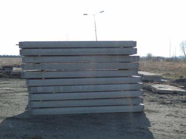 Stos prefabrykowanych płyt betonowych na placu budowy, widoczne uchwyty montażowe, w tle słupy oświetleniowe i pole. Jasne światło dzienne.