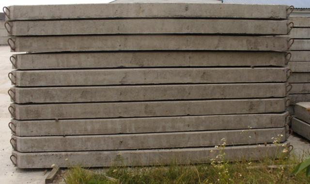 Stos prefabrykowanych płyt betonowych z metalowymi uchwytami, ułożonych równo na ziemi, z widoczną trawą i drobnymi kwiatami w dolnej części kadru.