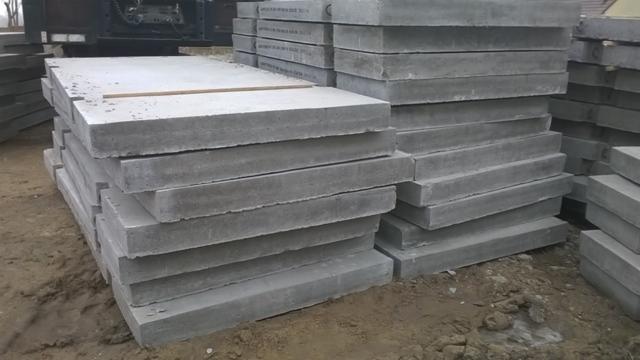 Stosy szarych, kwadratowych prefabrykatów betonowych ułożone na ziemi, z widocznym fragmentem maszyny w tle, prawdopodobnie na placu budowy.