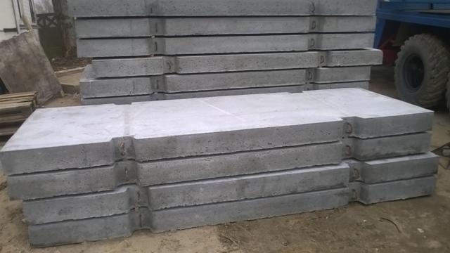 Stos prefabrykowanych, szarych płyt betonowych z uchwytami, ułożonych na ziemi. W tle widoczne koło samochodu ciężarowego i palety.