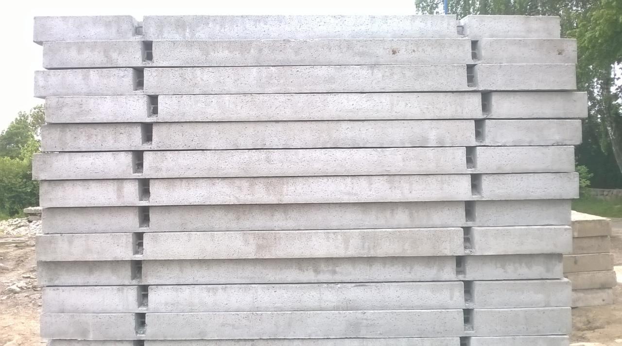 Stos prefabrykatów betonowych z charakterystycznymi wycięciami, ułożonych jeden na drugim, w tle zieleń drzew. Szary beton kontrastuje z naturalnym otoczeniem.