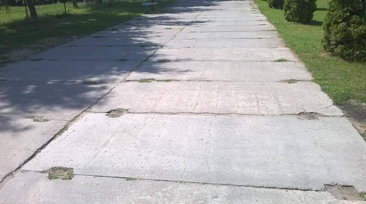 Betonowa droga z widocznymi pęknięciami i porastającą trawą, otoczona zielenią i drzewami. Fragment chodnika z prefabrykatów betonowych na terenie zielonym.