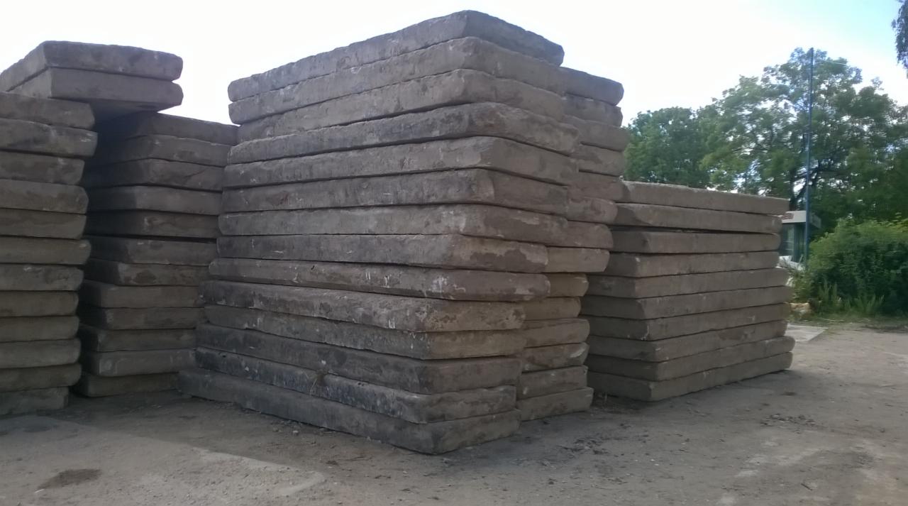 Stosy szarych, betonowych płyt chodnikowych ułożone na ziemi, widoczne na tle zieleni drzew. Płyty o zaokrąglonych krawędziach.