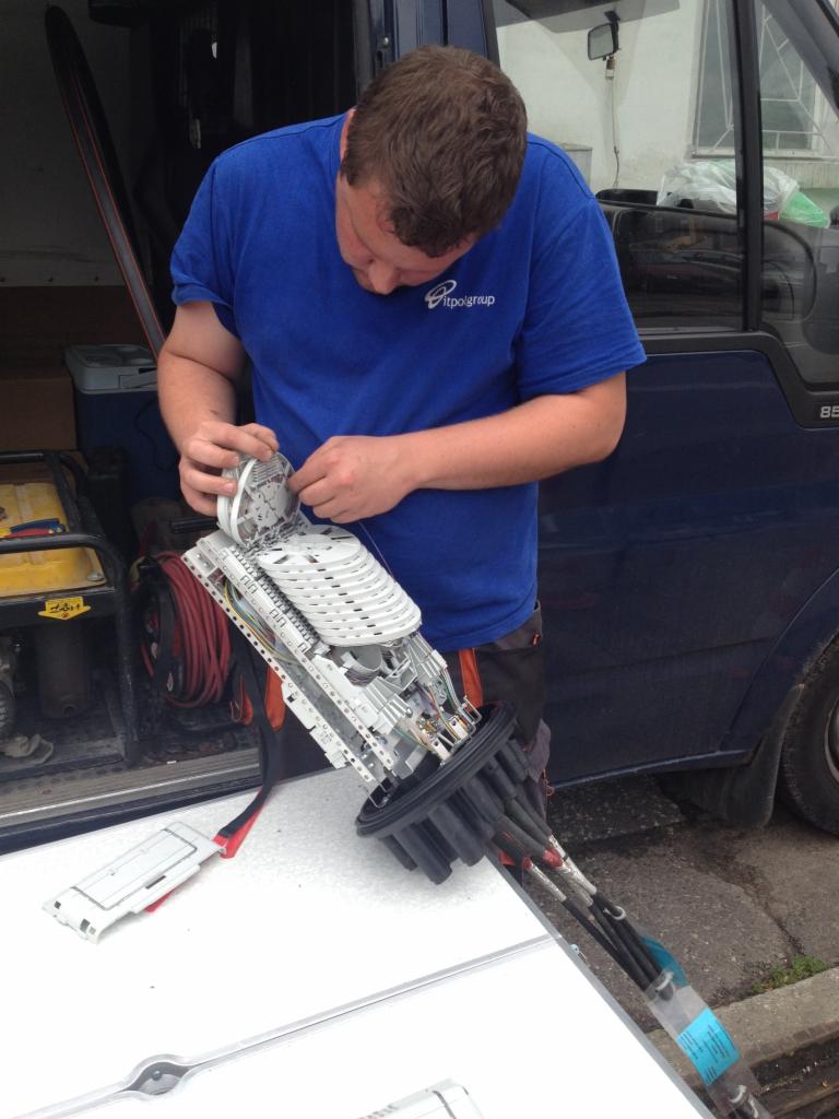 Pracownik w niebieskiej koszulce z logo firmy łączy kable światłowodowe w mufie na prowizorycznym stole na tle samochodu dostawczego.