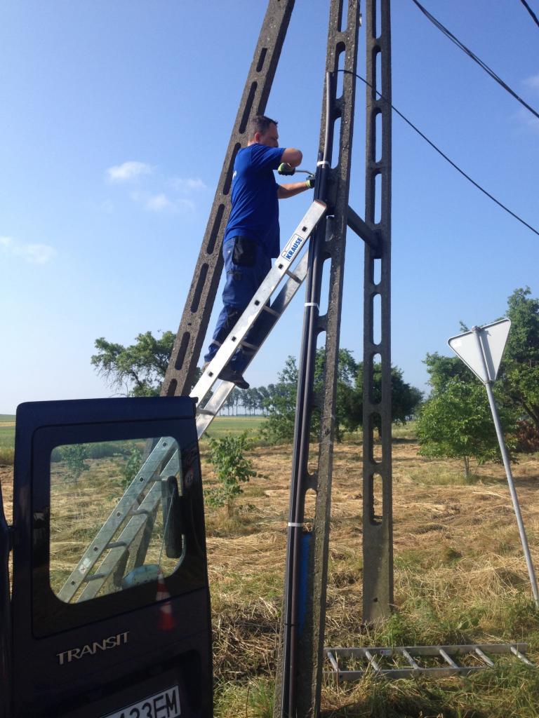 Pracownik na drabinie, mocuje przewody na betonowym słupie telekomunikacyjnym, widoczny samochód dostawczy w tle.