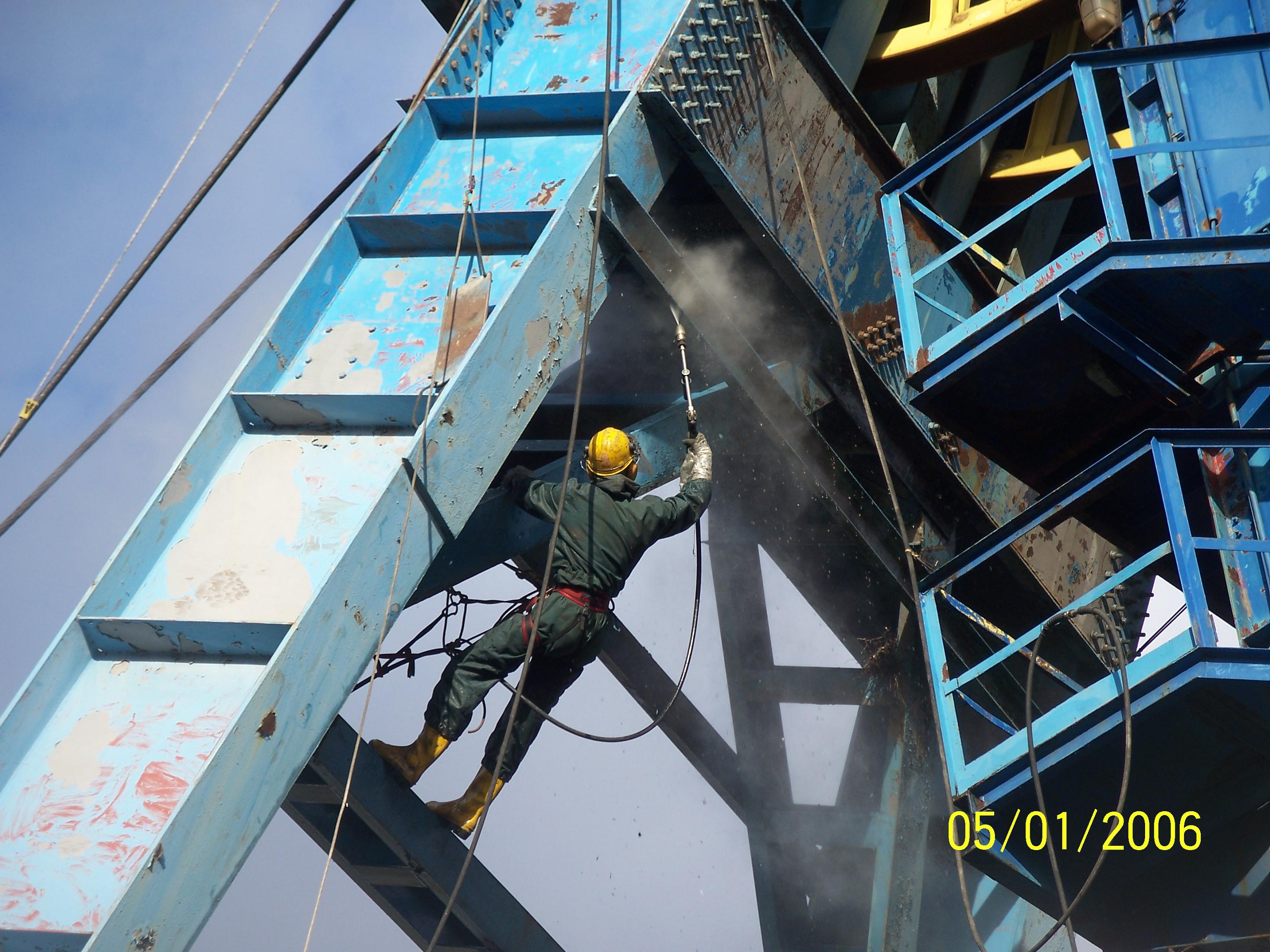 Pracownik w kasku i kombinezonie, zabezpieczony linami, czyści metalową konstrukcję metodą strumieniowo-ścierną, widoczne odpryski rdzy i farby.