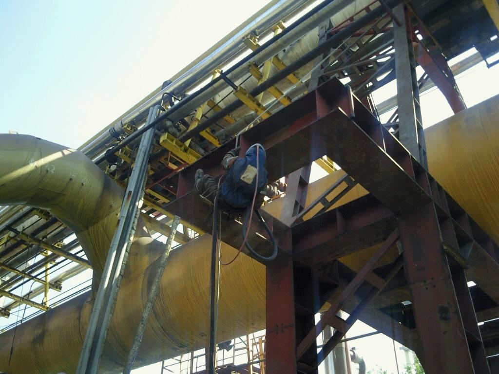 Pracownik w kombinezonie, zabezpieczony linami, wykonuje prace na wysokości na konstrukcji stalowej z rurociągami. Widoczne narzędzia i przewody. Rurociągi pokryte żółtą izolacją.