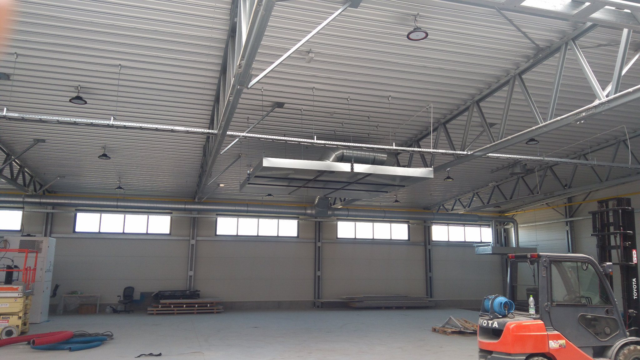 Wnętrze hali produkcyjnej z zamontowaną, podwieszaną instalacją wentylacyjną z widocznymi kanałami wentylacyjnymi, oświetleniem i konstrukcją dachu z blachy trapezowej oraz wózkiem widłowym w tle.
