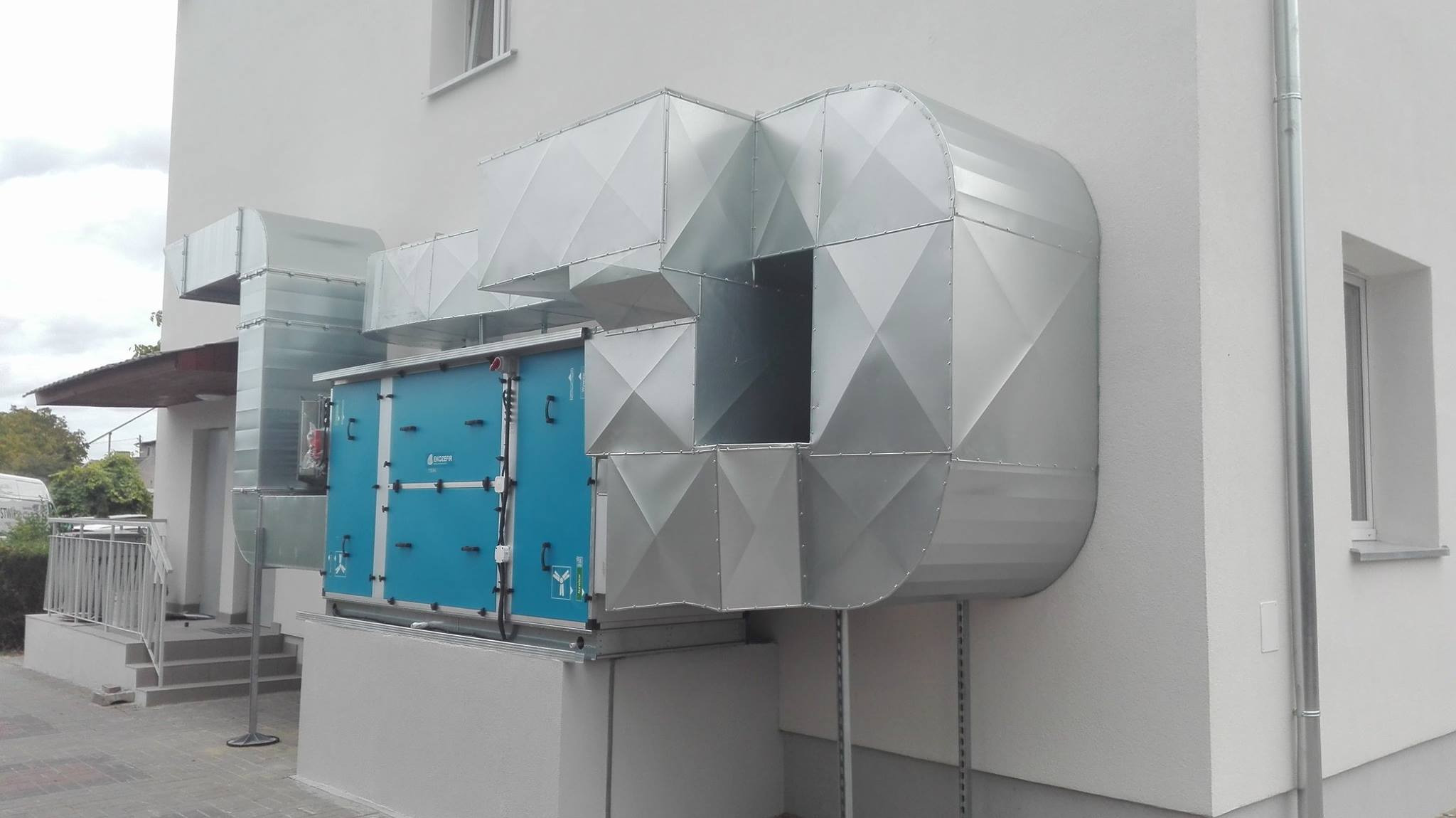 Zewnętrzna instalacja wentylacyjna z centralą w niebieskiej obudowie i srebrnymi kanałami wentylacyjnymi przymocowanymi do ściany budynku.