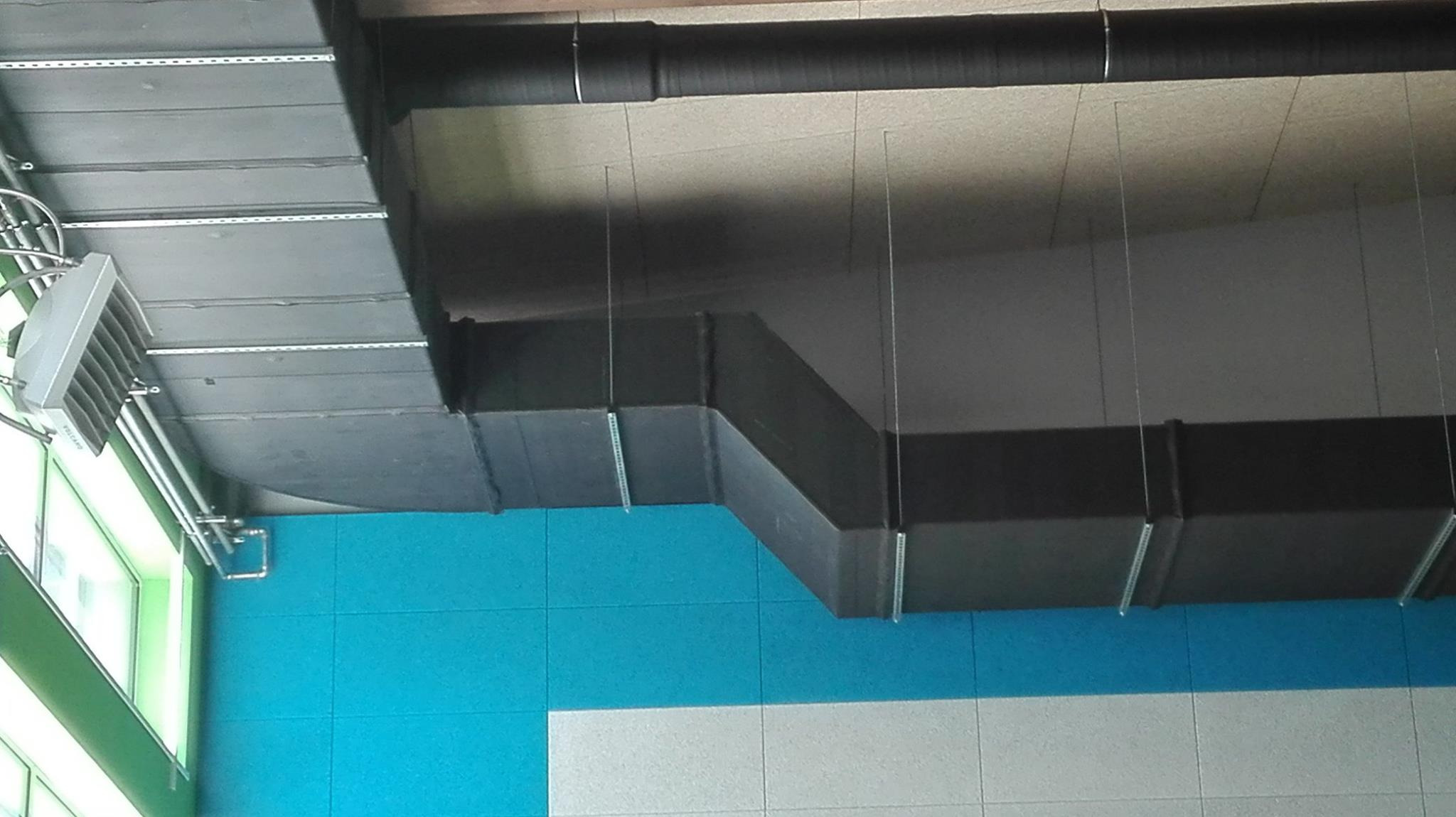 Szare kanały wentylacyjne prostokątne podwieszone pod sufitem, widoczne elementy montażowe i niebiesko-biała ściana w tle, lampa przemysłowa przy oknie.