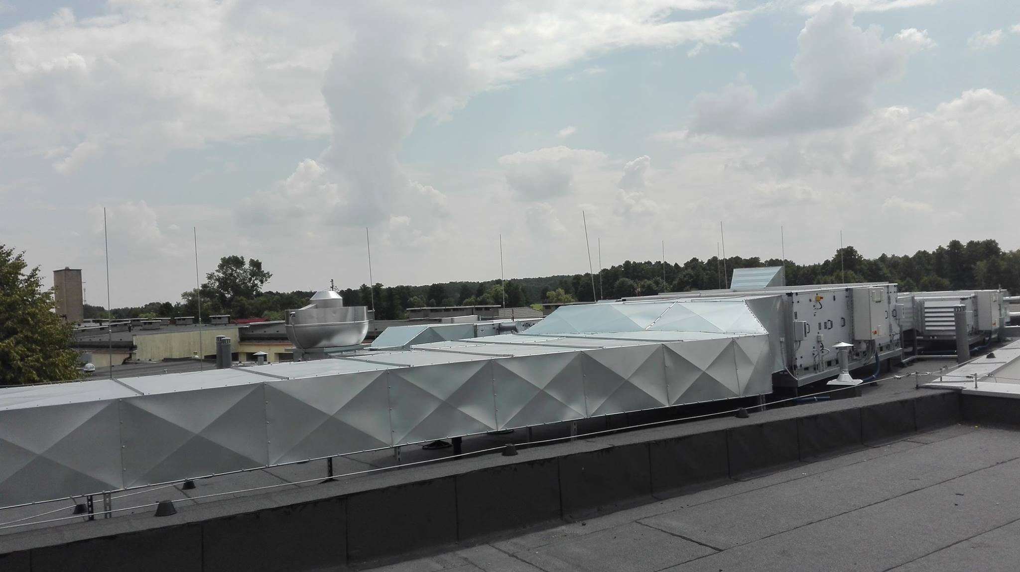 System kanałów wentylacyjnych z widocznymi elementami centrali wentylacyjnej na dachu budynku, z antenami odgromowymi i drzewami w tle pod pochmurnym niebem.