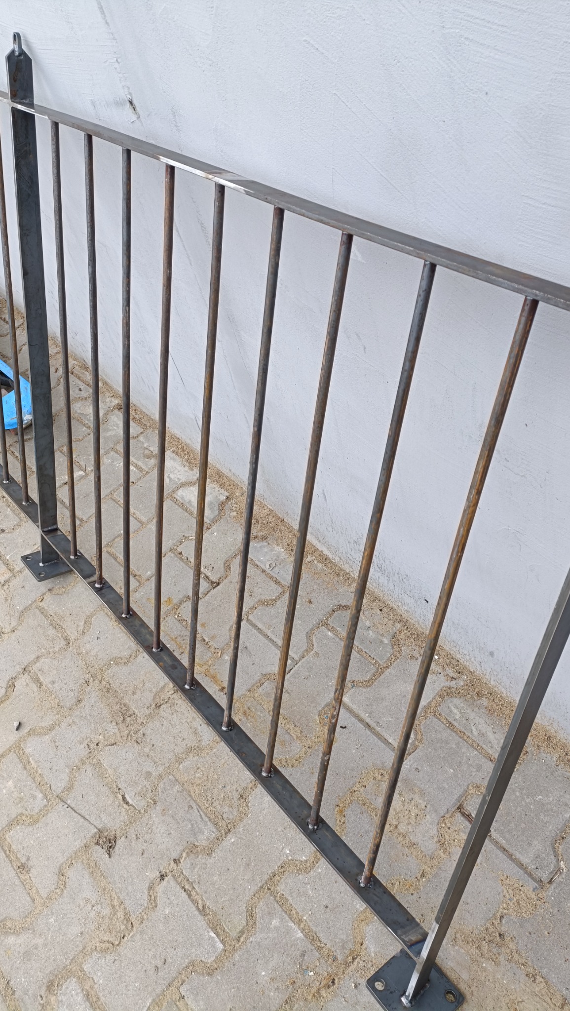 Surowy, metalowy fragment ogrodzenia z pionowymi prętami, przyspawany do poziomej belki i mocowany do podłoża za pomocą metalowych stopek, na tle białej ściany i betonowej kostki brukowej.