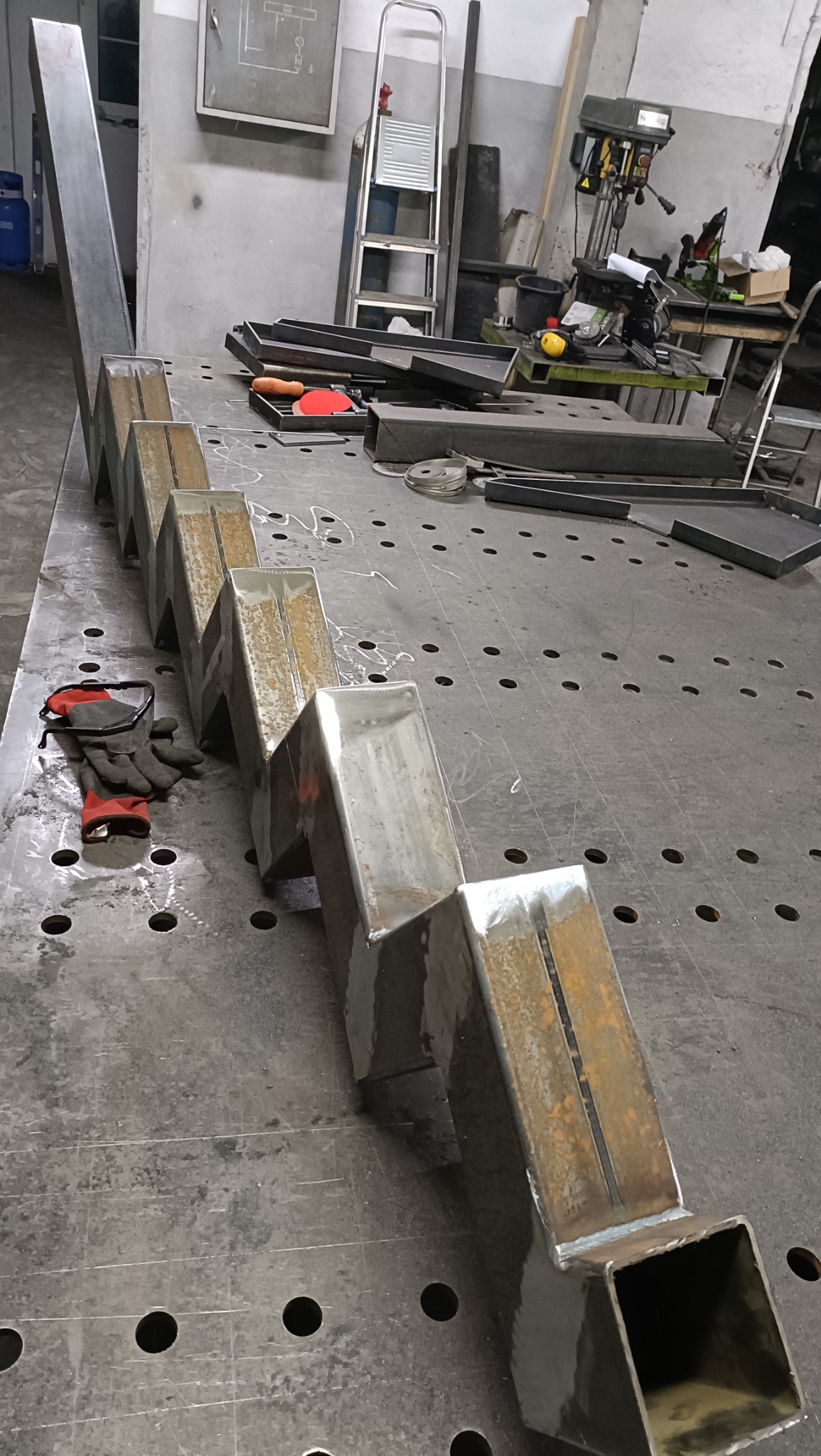 Metalowa konstrukcja schodkowa spawana z profili kwadratowych, leżąca na perforowanym stole warsztatowym, obok rękawice robocze; w tle drabina i wiertarka stołowa.