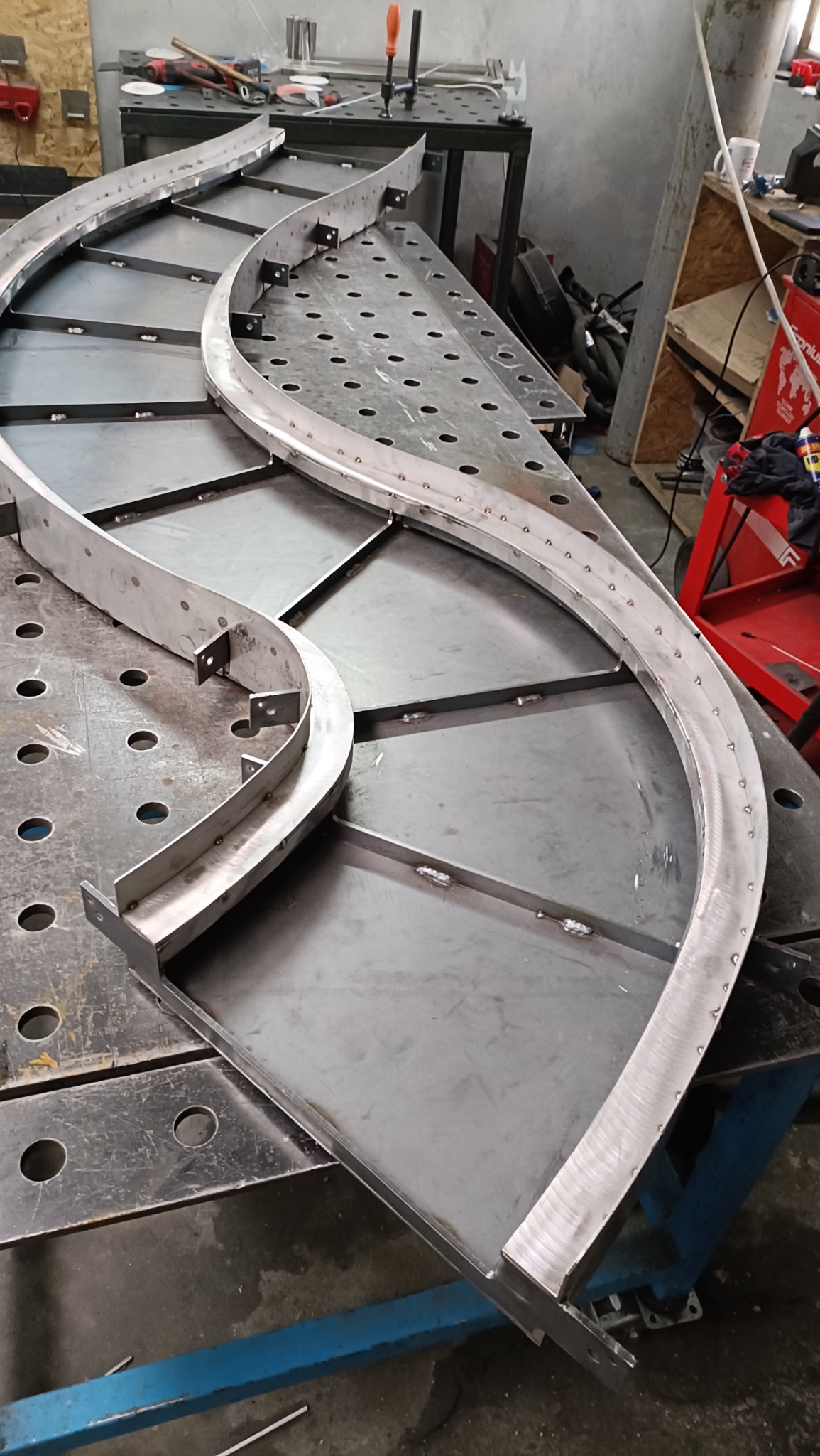 Metalowa konstrukcja z wygiętymi elementami, prawdopodobnie fragment większej całości, spawana na stole warsztatowym z widocznymi śladami obróbki.