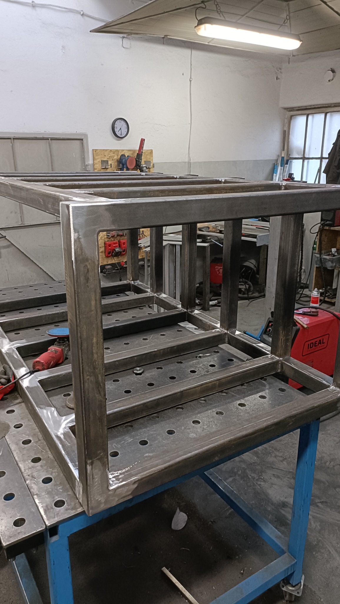 Spawana, metalowa konstrukcja szkieletowa, prawdopodobnie stelaż meblowy, na perforowanym stole spawalniczym w warsztacie, widoczny spawany narożnik. W tle spawarka.