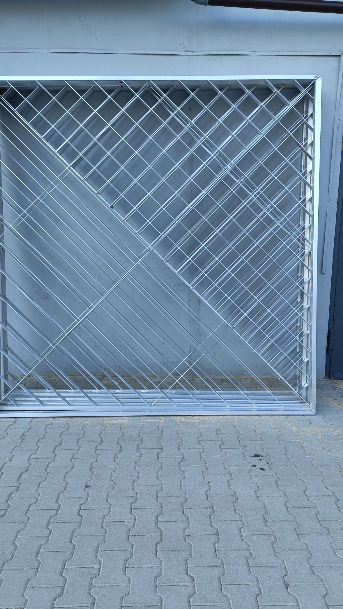 Metalowa rama z kratownicą o ukośnym wzorze, oparta o szarą ścianę, ułożona na chodniku z betonowej kostki.