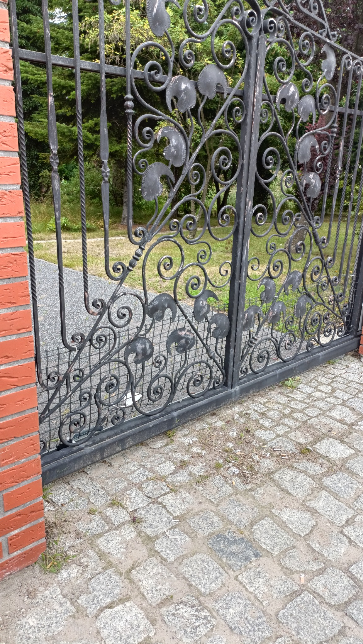 Metalowa brama z ozdobnymi elementami w kształcie liści, zamontowana na ceglanym słupku, widok na kostkę brukową i zieleń w tle.