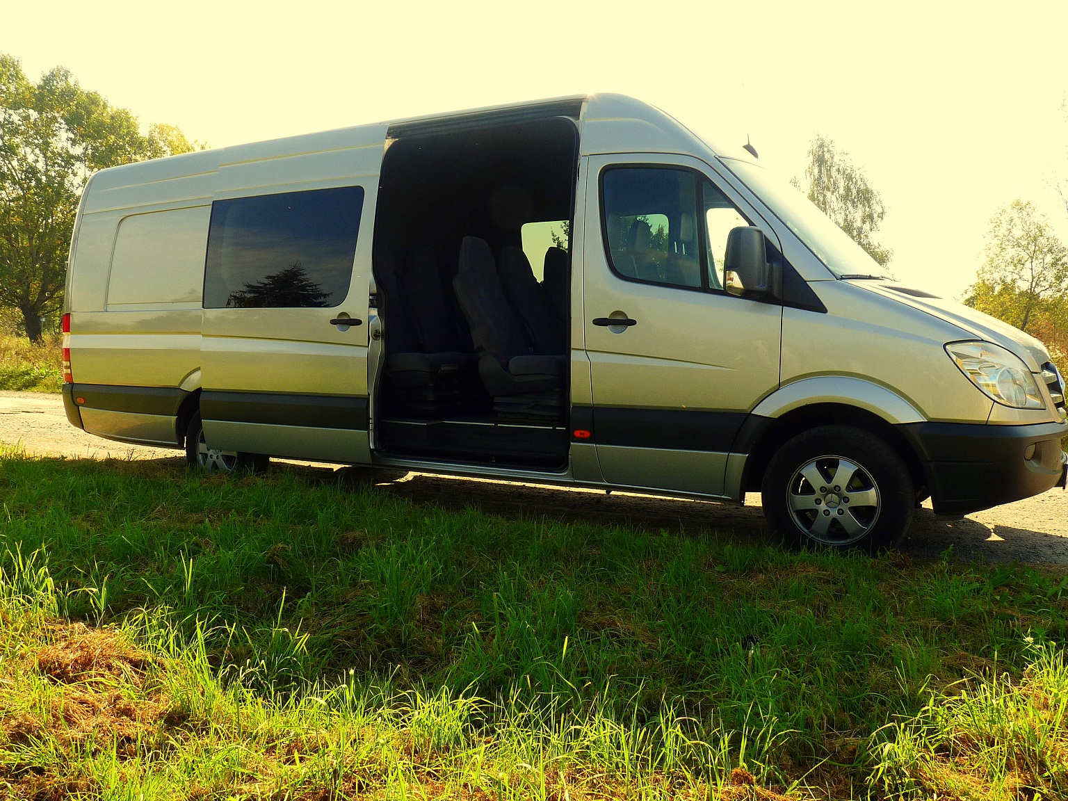 Srebrny bus z otwartymi drzwiami przesuwnymi, zaparkowany na trawiastym poboczu drogi, widoczne fotele wewnątrz.