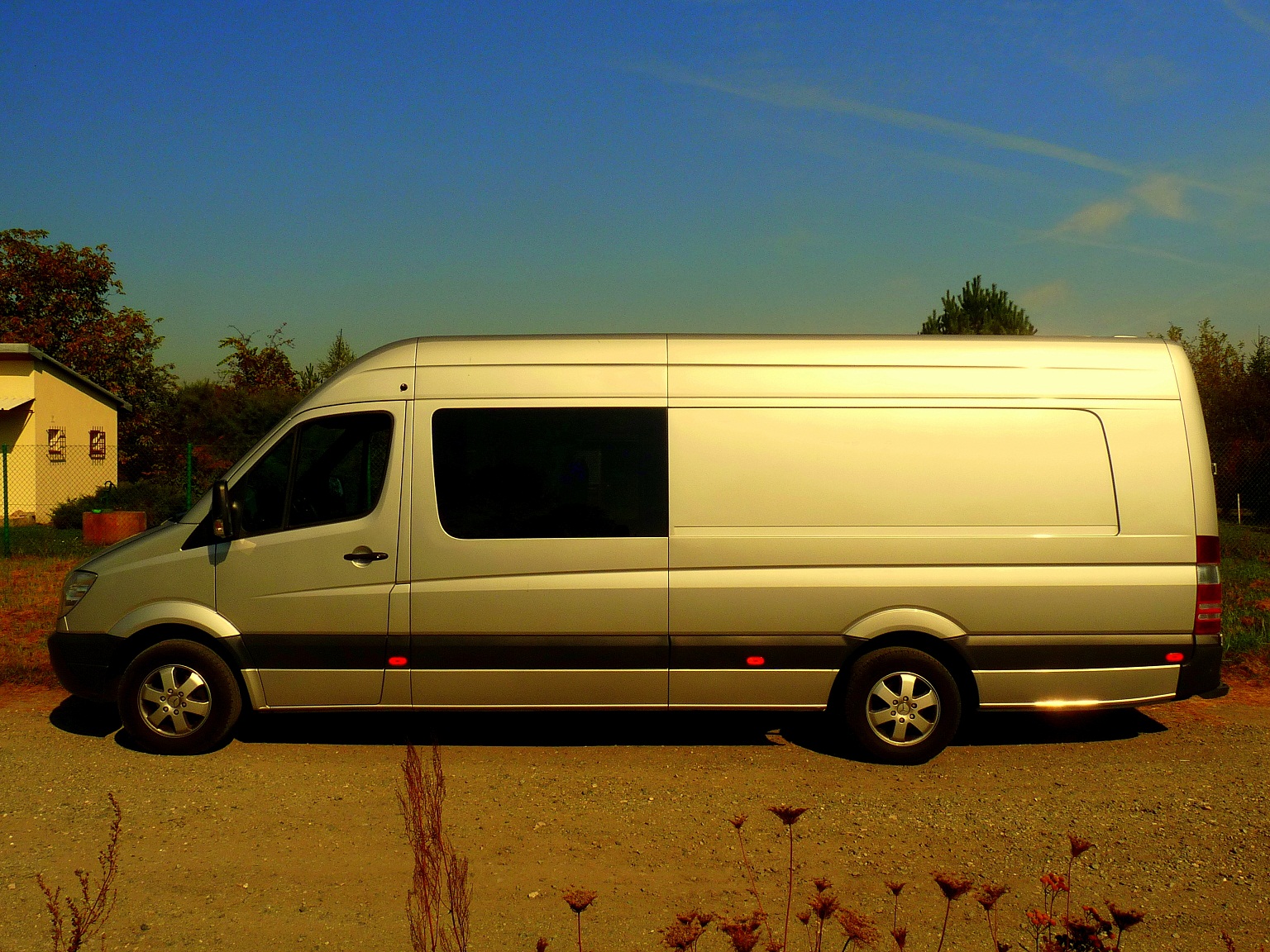 Srebrny bus osobowo-towarowy Mercedes-Benz Sprinter zaparkowany na żwirowej drodze w słoneczny dzień, widok z boku, z widocznym budynkiem gospodarczym w tle.