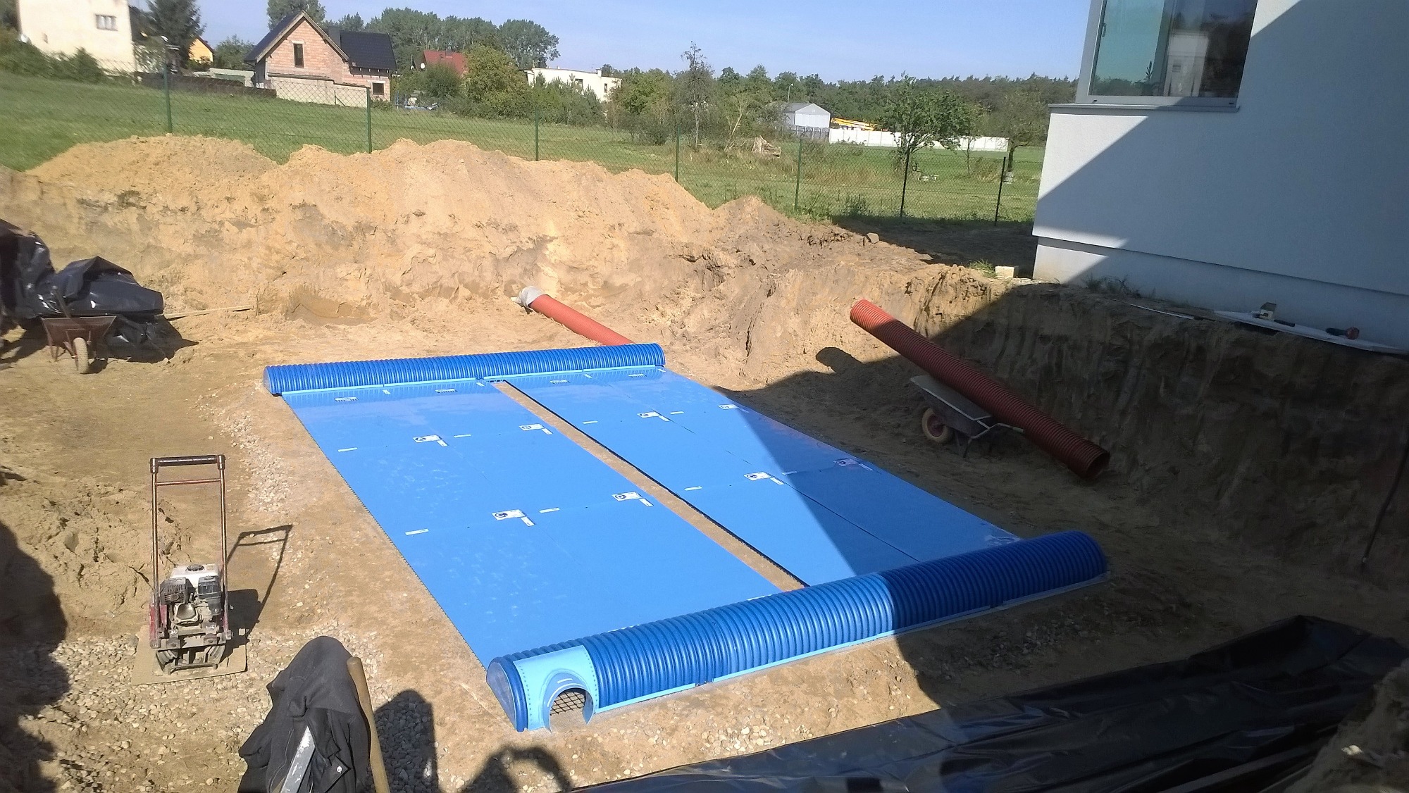 Wykonana instalacja gruntowego wymiennika ciepła (GWC) w wykopie, z widocznymi rurami karbowanymi i niebieską folią izolacyjną, zagęszczarka w tle.