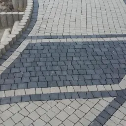 Ułożona kostka brukowa w dwóch odcieniach szarości, tworząca wzór na podjeździe, z obrzeżami z betonowych elementów w kształcie walców, częściowo jeszcze nieukończonymi.