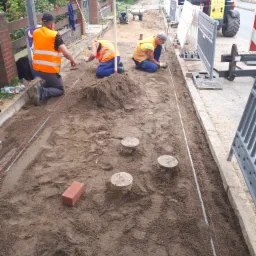 Trzech robotników w odblaskowych kamizelkach klęczy na ziemi, układając kable lub rury w wykopie wzdłuż chodnika, widoczny sprzęt budowlany i znak drogowy w tle.