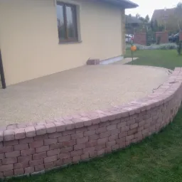 Wykończony taras z ceglanym murkiem przy domu z beżową elewacją. Widoczna trawa i elementy ogrodu w tle. Nowoczesne rozwiązanie.