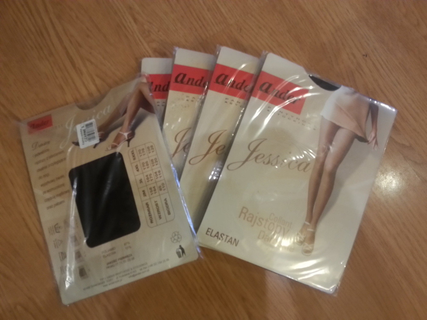 Kilka opakowań rajstop marki Jessica leżących na drewnianej powierzchni, jedno opakowanie czarnych rajstop z widoczną tabelą rozmiarów, pozostałe opakowania w odcieniach beżu z grafiką nóg w butach...