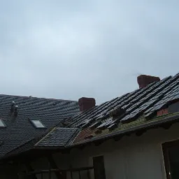 Dwa dachy pokryte dachówką, jeden w trakcie układania z widocznymi łatami, drugi z oknami dachowymi i kominem, pochmurne niebo w tle.