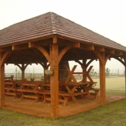 Drewniana altana z ciemnoczerwoną dachówką, z solidną konstrukcją wspartą na zdobionych kolumnach, wyposażona w stoły i ławy, otoczona brukowanym placem i trawnikiem pokrytym szronem w mglisty...