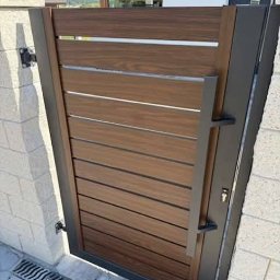 AAR PW Szczęsne mgr inż. Stefan Ruchlewicz - Nowoczesna furtka z poziomych desek w odcieniach brązu, z metalową ramą i długim uchwytem, zamontowana na murowanym słupku z jasnej cegły.