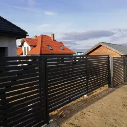 Nowoczesne, poziome, czarne ogrodzenie lamelowe na tle domów z dachówką i budynku gospodarczego, widoczne ślady prac ziemnych.